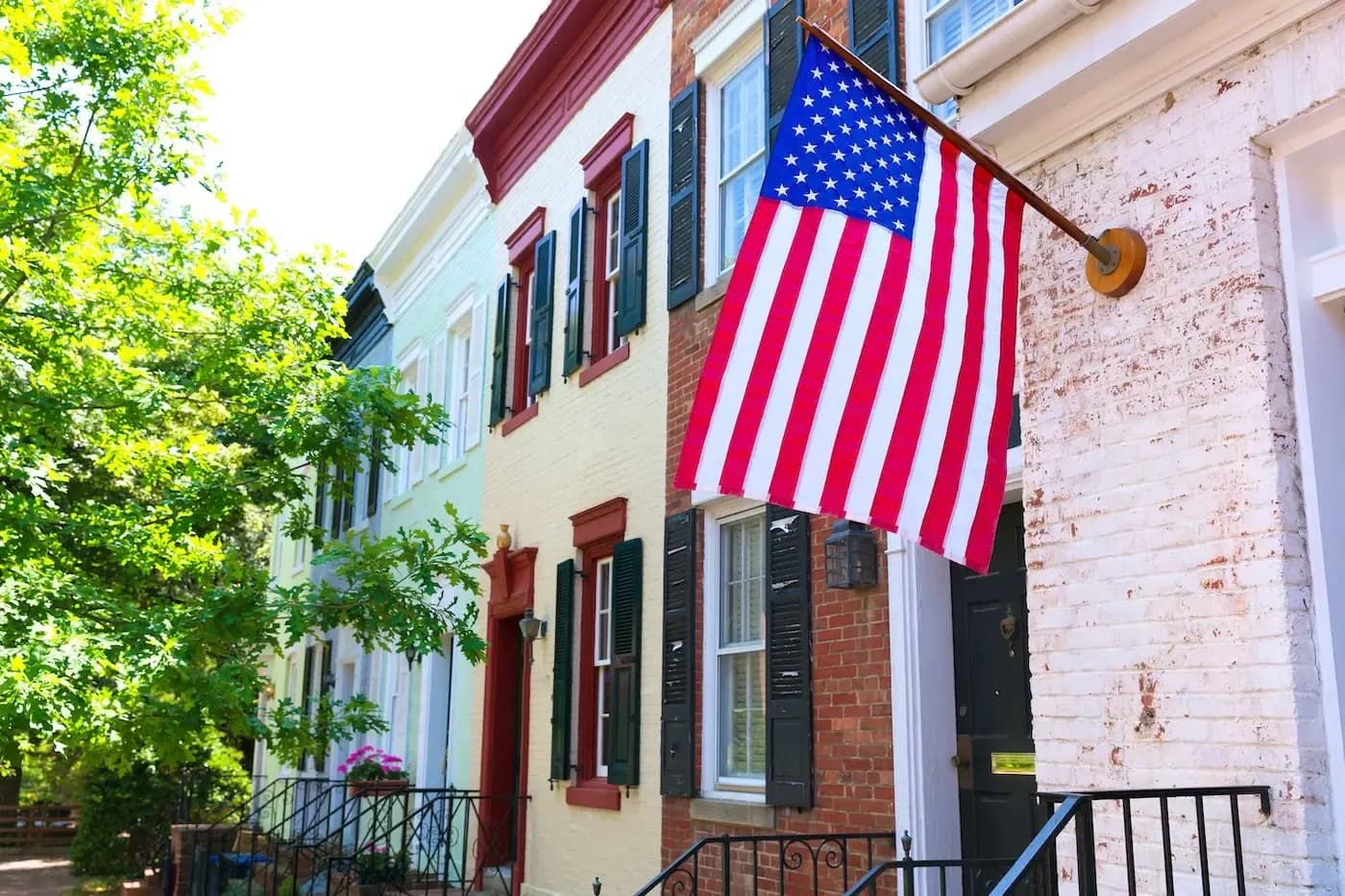 Gran bandera estadounidense colgada en un edificio histórico de ladrillo, con casas adosadas de colores en una calle arbolada