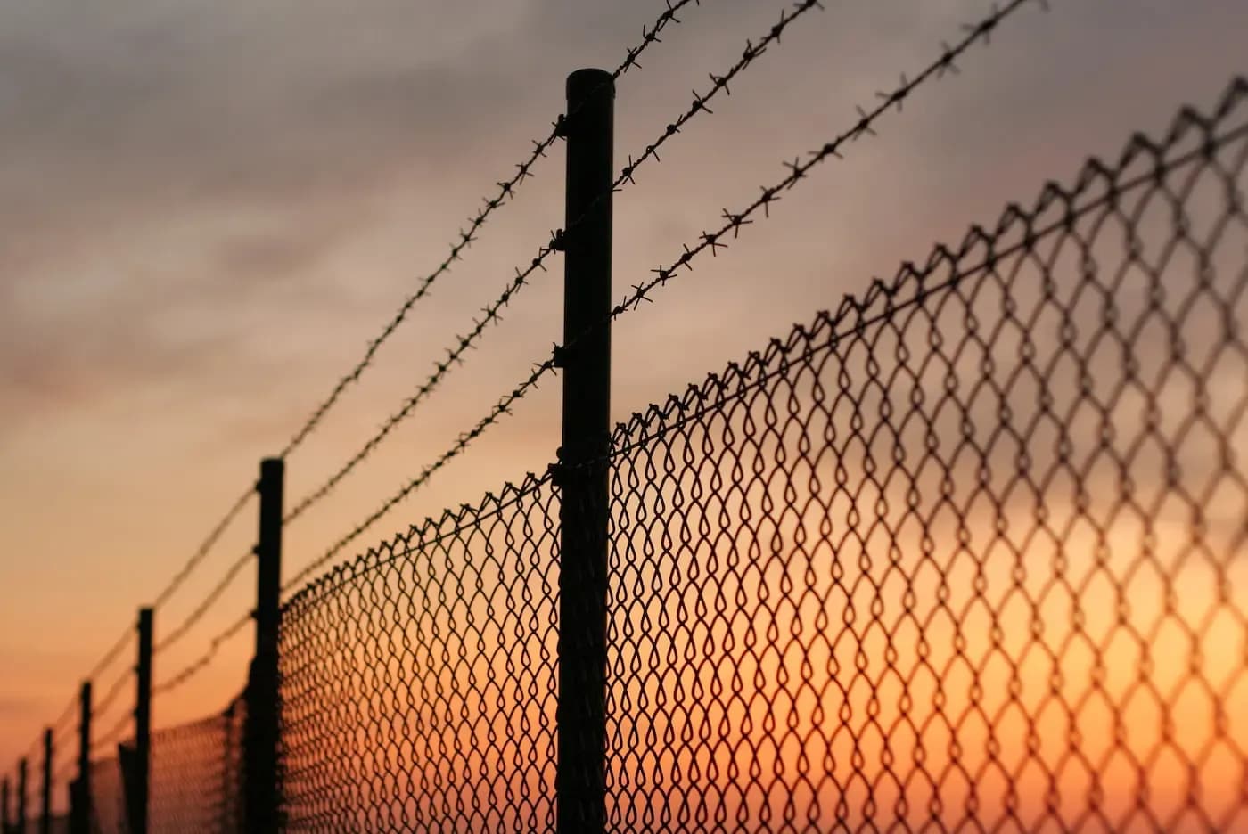 Silhouetted barbed wire fence and chain-link barrier against an orange and purple sunset sky