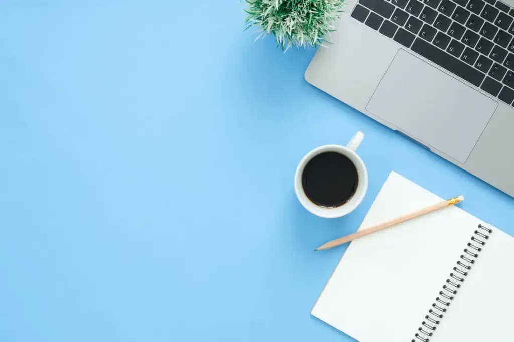 Laptop, coffee cup, and spiral notebook with pencil on blue desk