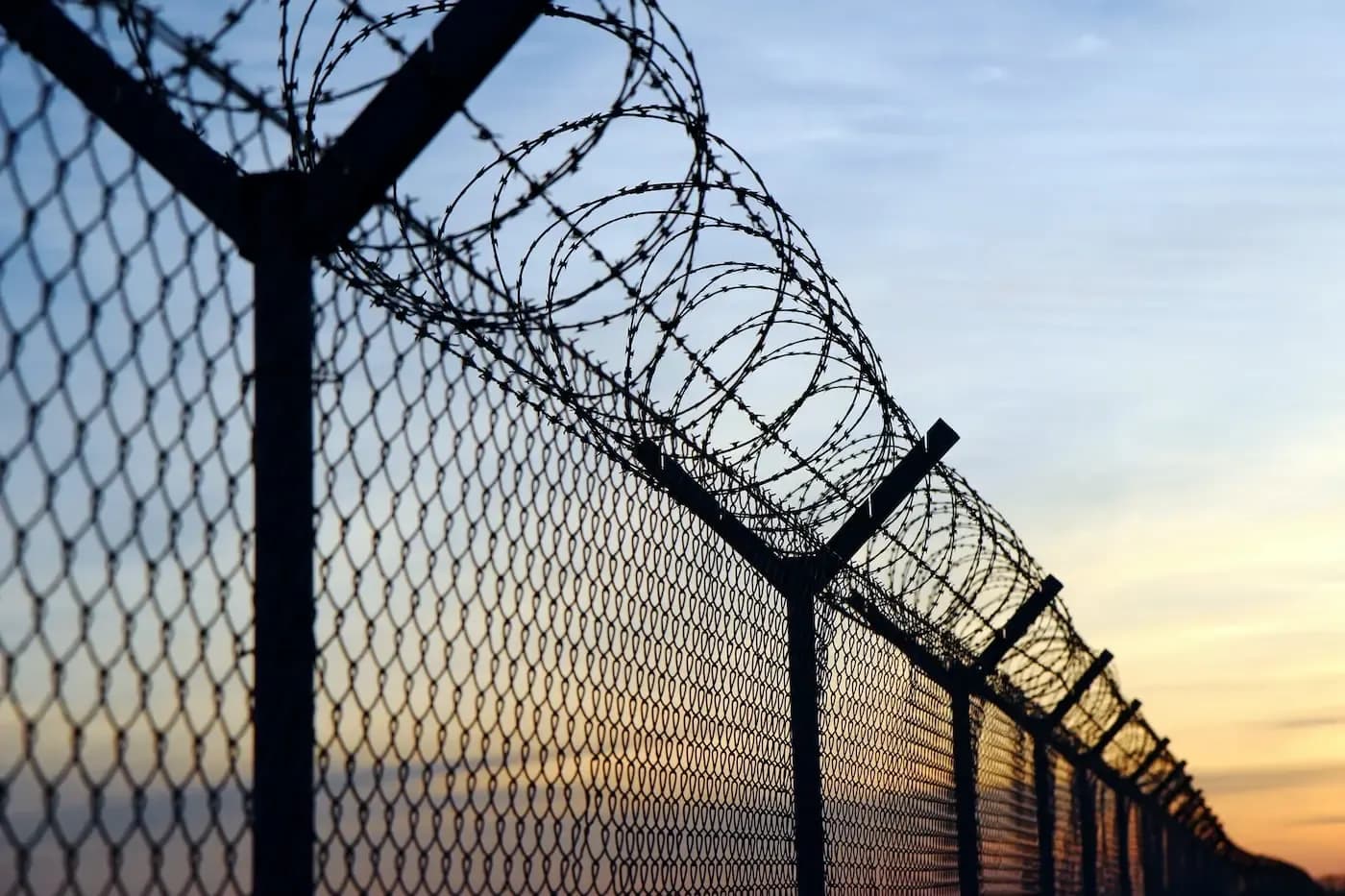 Razor wire atop a chain-link fence silhouetted against a sunset sky
