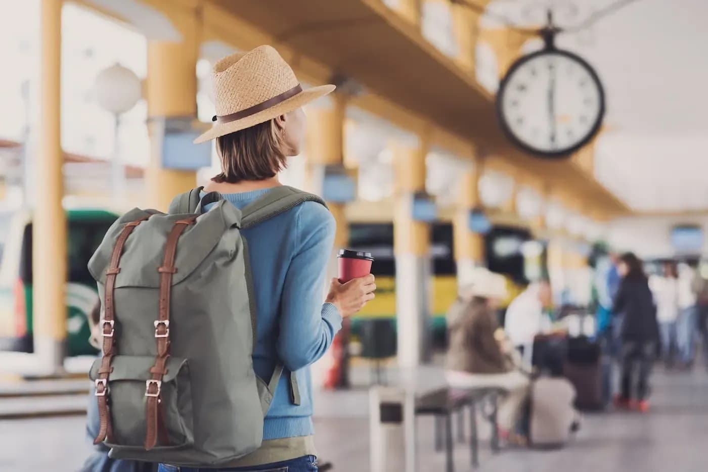Persona con mochila y sombrero sosteniendo una taza de café en el andén de una concurrida estación de transporte