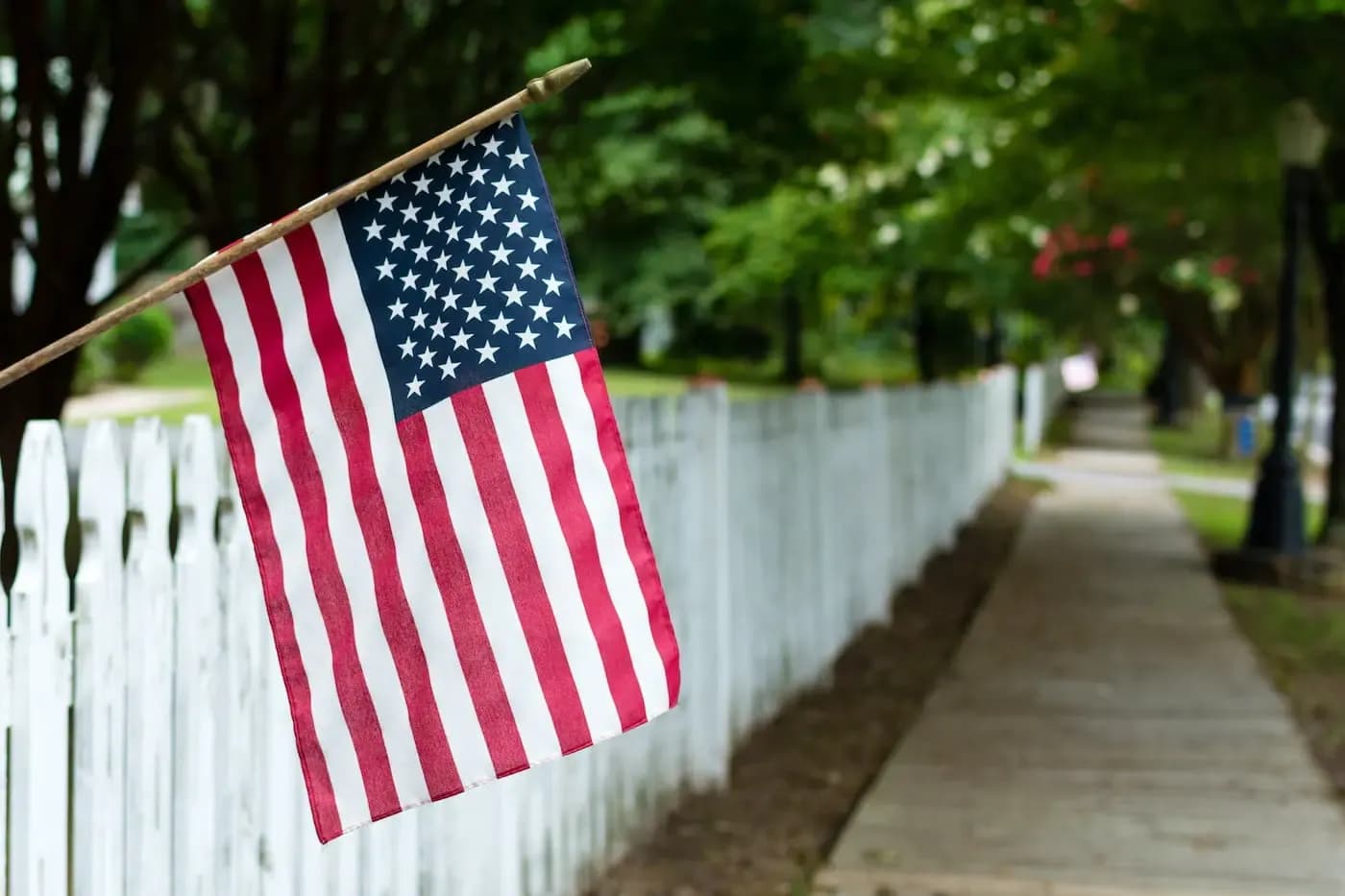 American flag on white picket fence along cemetery pathway with green trees