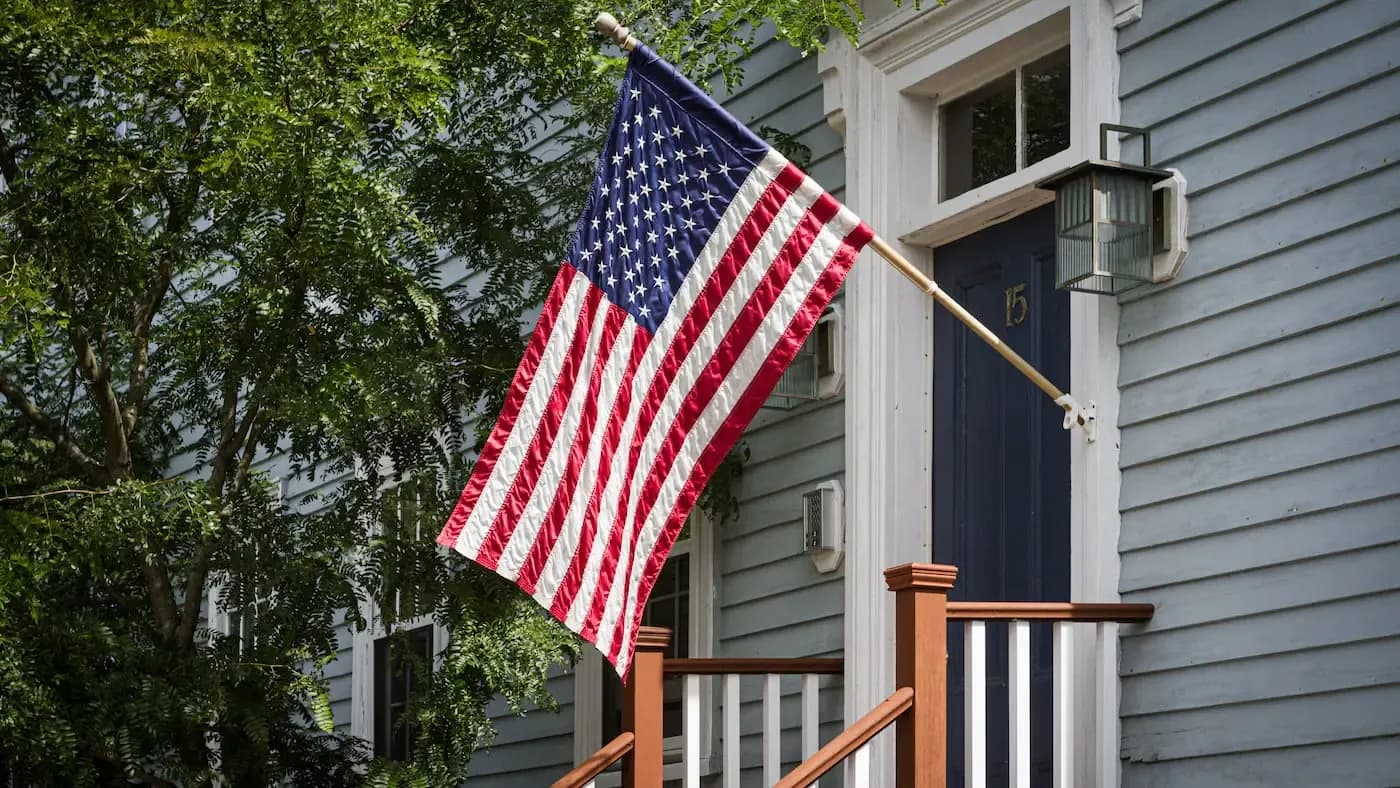 American flag hanging on wooden bracket of a traditional colonial-style house with green shutters