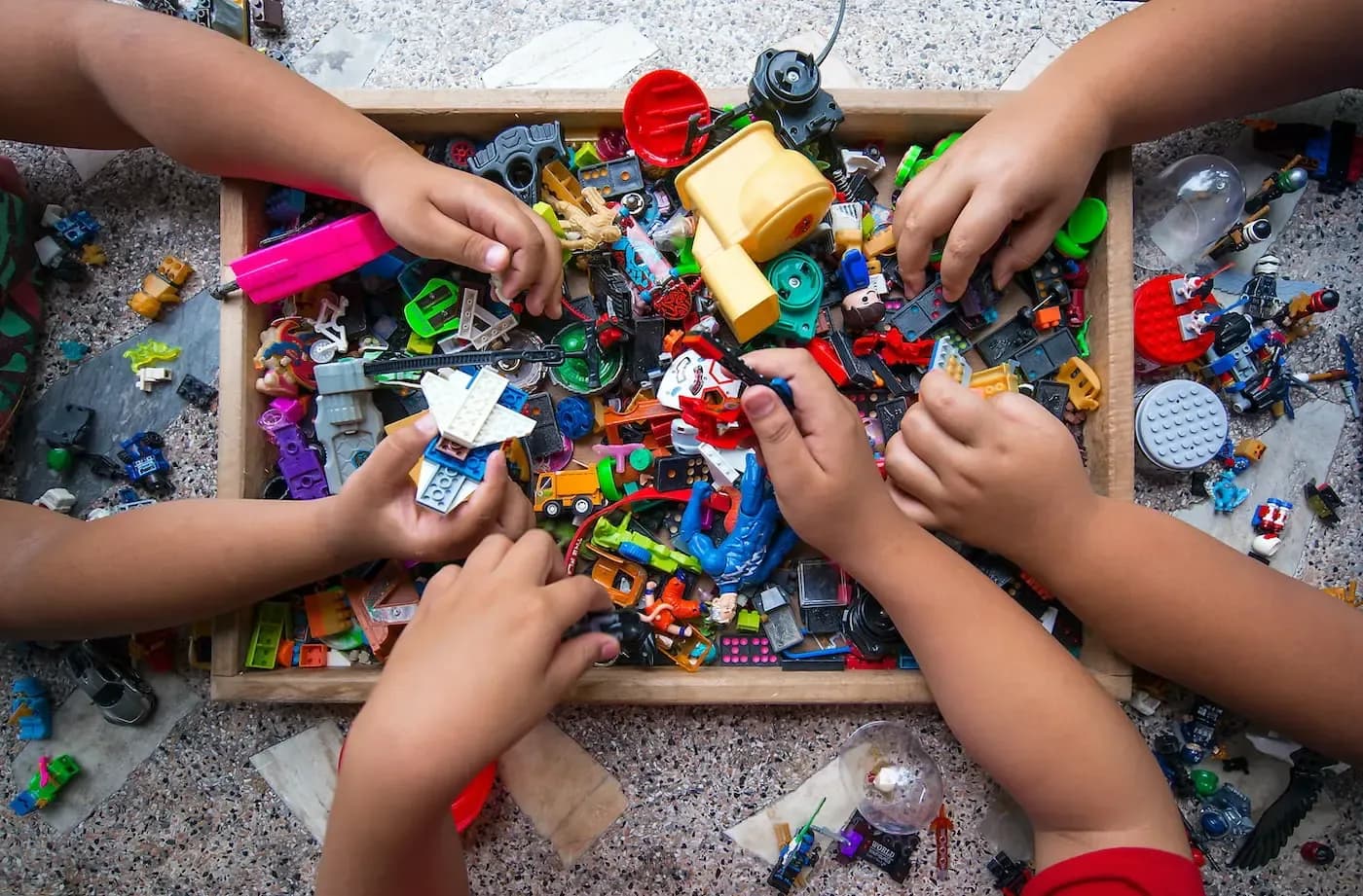 Multiple children's hands reaching into a wooden box filled with colorful toys and building blocks