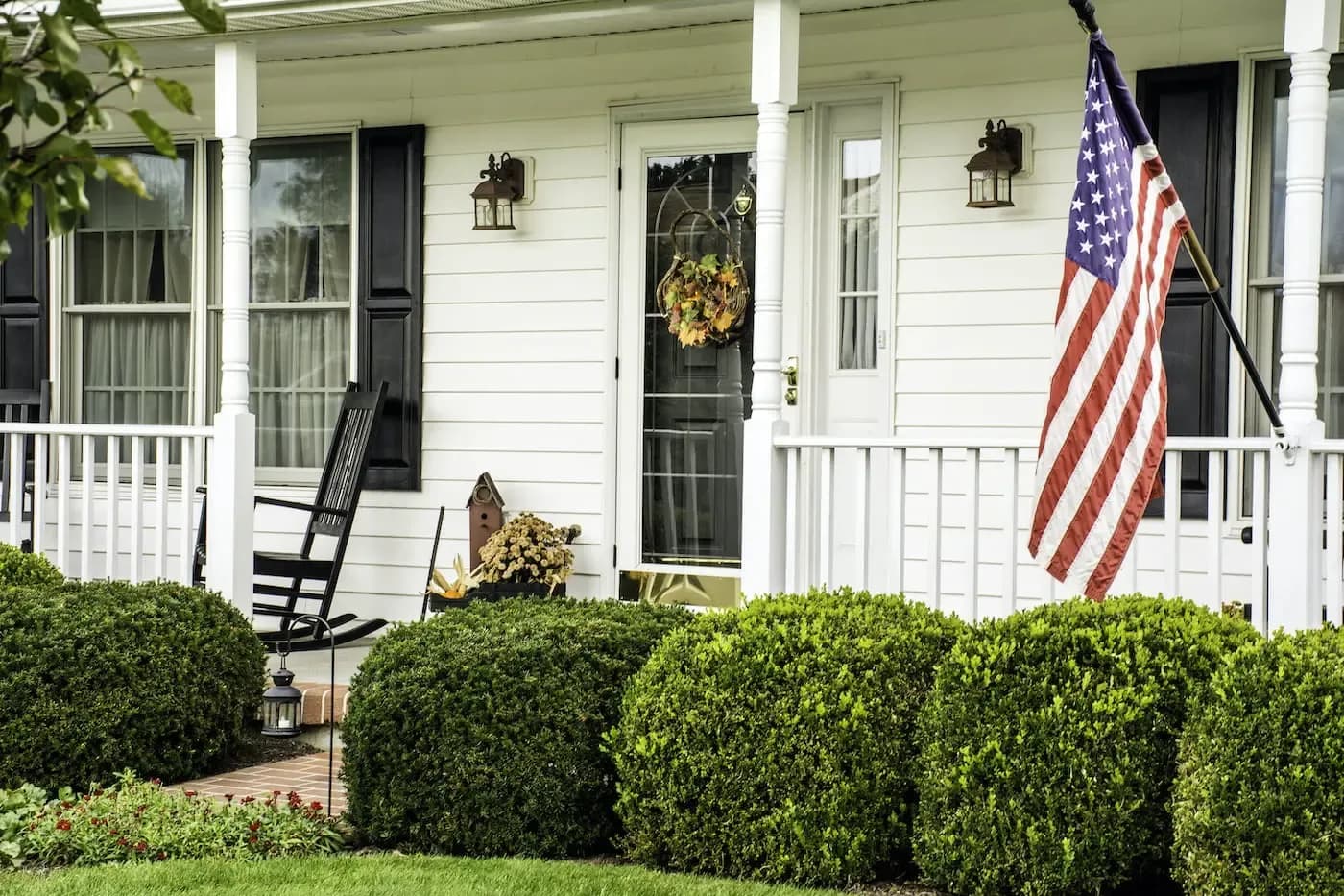 White columned porch with American flag, rocking chair, and manicured hedges