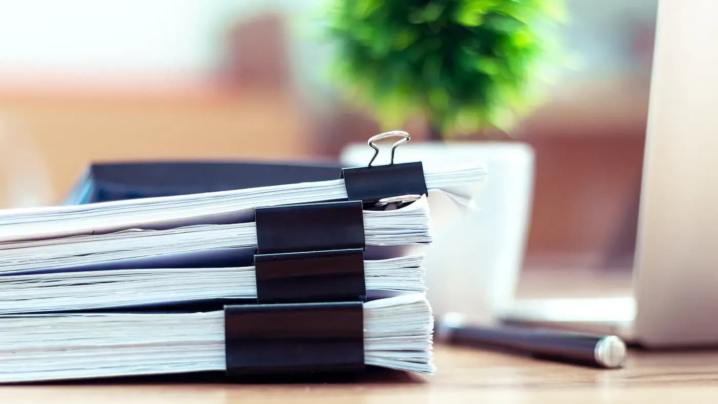 Stack of organized documents with black binder clips and a potted plant in the background on a desk