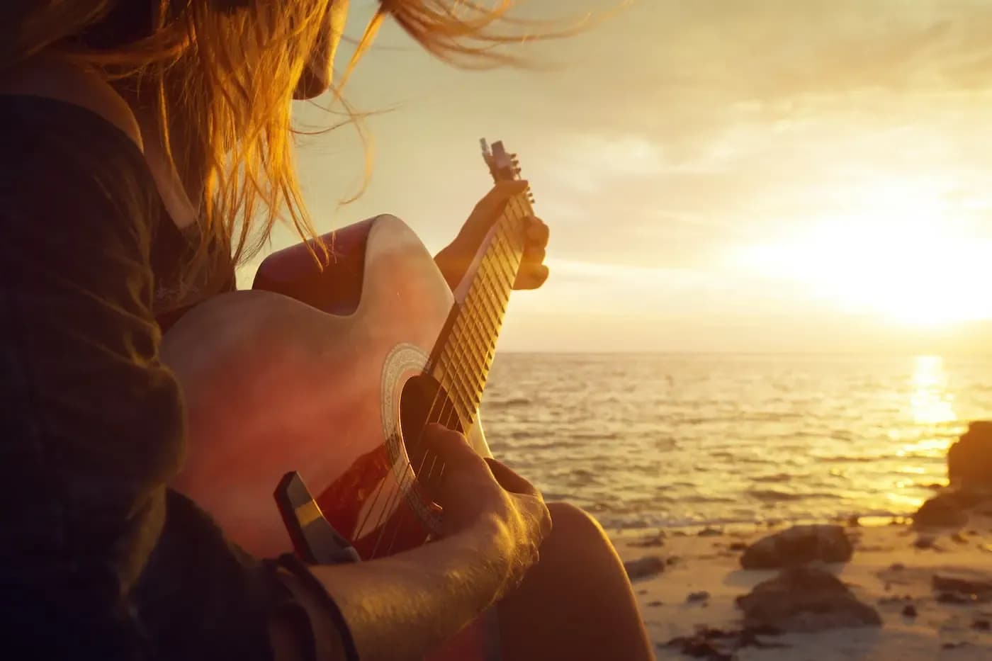 Person playing acoustic guitar on beach at golden sunset over ocean