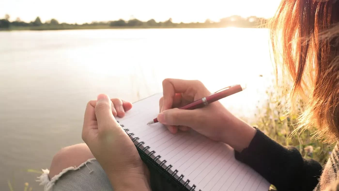 Person with red hair writing in a notebook beside a calm lake at sunset